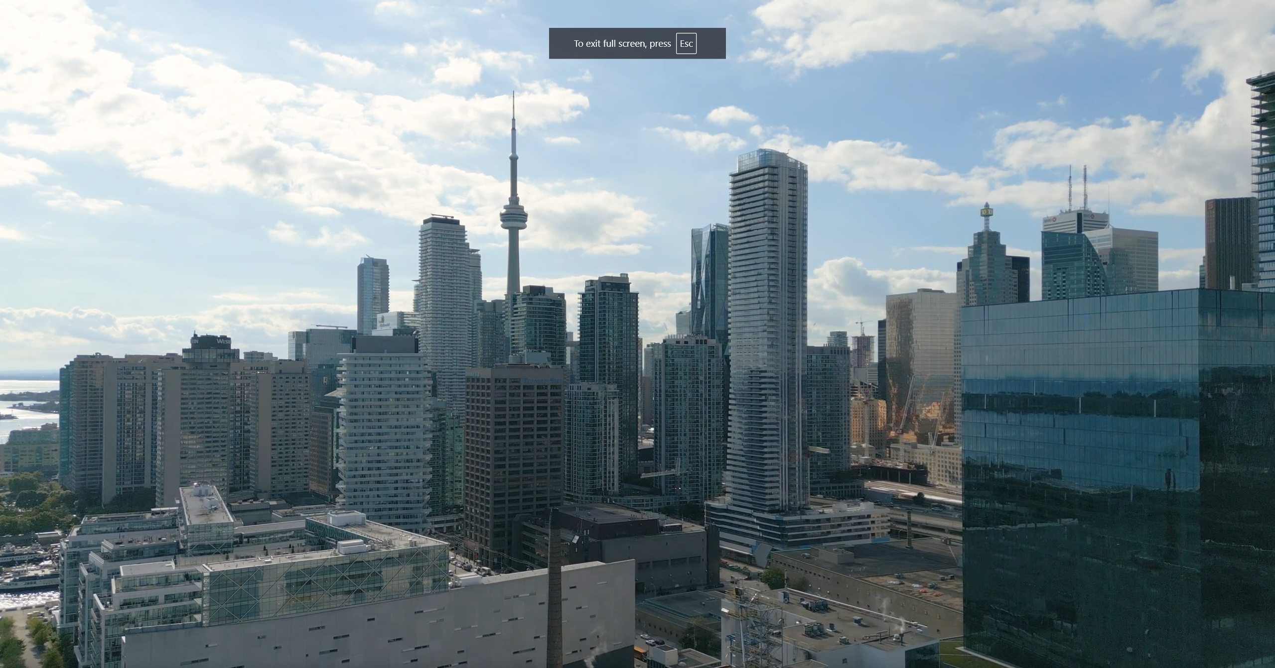 Aerial view of downtown Toronto skyline featuring the iconic CN Tower and modern glass skyscrapers against a cloudy blue sky, with Lake Ontario visible in the background